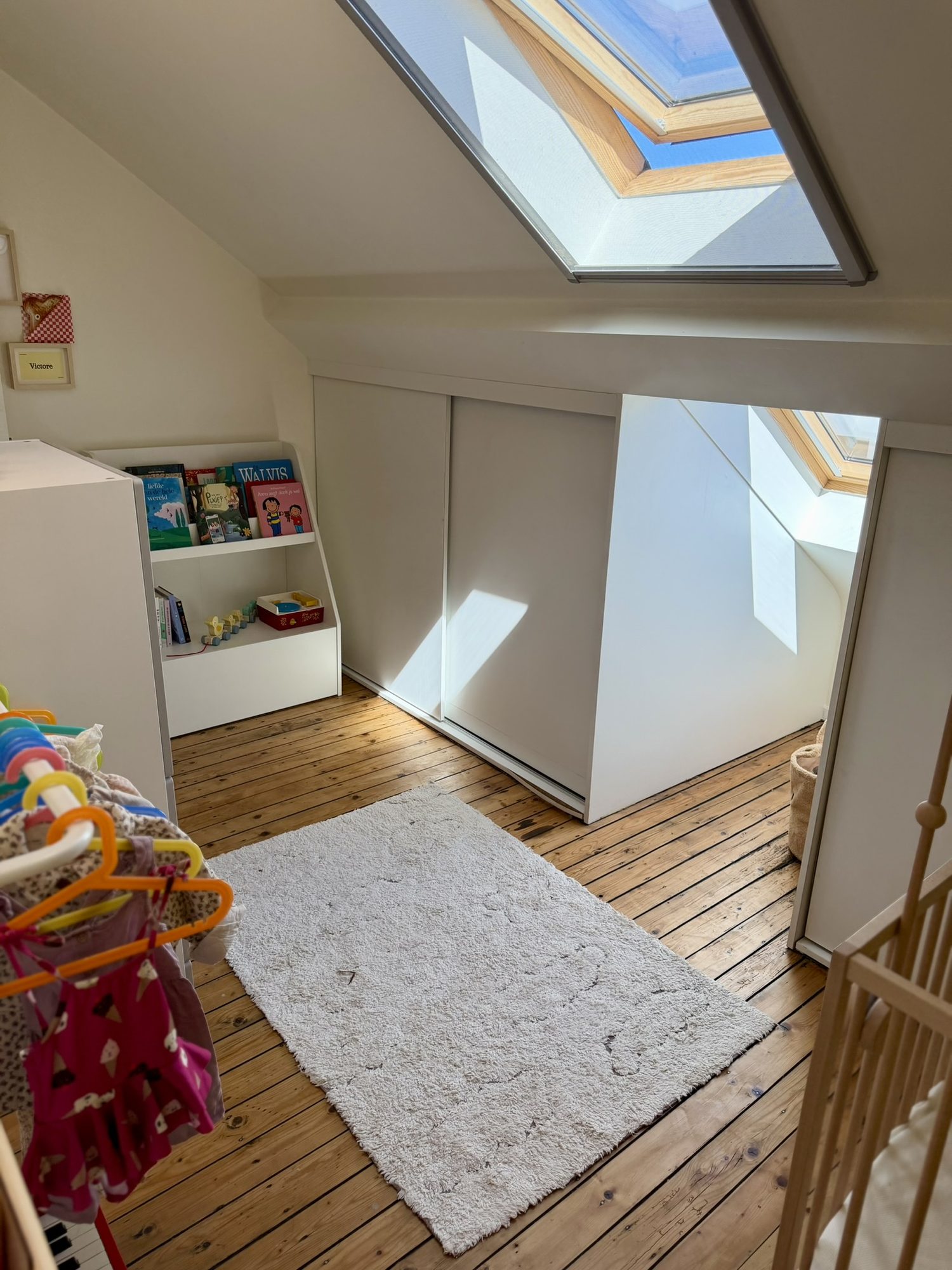 Zonnige kinderkamer onder het dakraam met massieve houten vloer, leeshoek, speelgoed en doorkijk naar de aanpalende ruimte